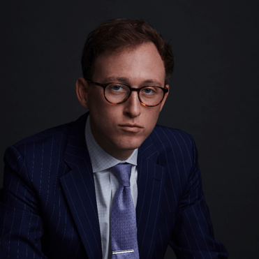 A man wearing glasses, a blue pinstripe suit, a light blue shirt, and a lavender tie sits against a dark background, looking straight at the camera with a serious expression.