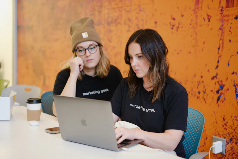 WMLF0720 Two women wearing marketing geek t-shirts sit at a white table, focused on a laptop. One wears glasses and a beanie, the other types. A coffee cup and takeout container are nearby. The background is a textured orange wall.
