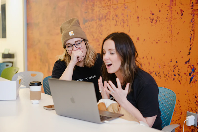 Two women sit at a table looking at a laptop. One wears glasses and a beanie, while the other gestures animatedly with her hands. Both appear engaged and are wearing black t-shirts. A coffee cup sits on the table.